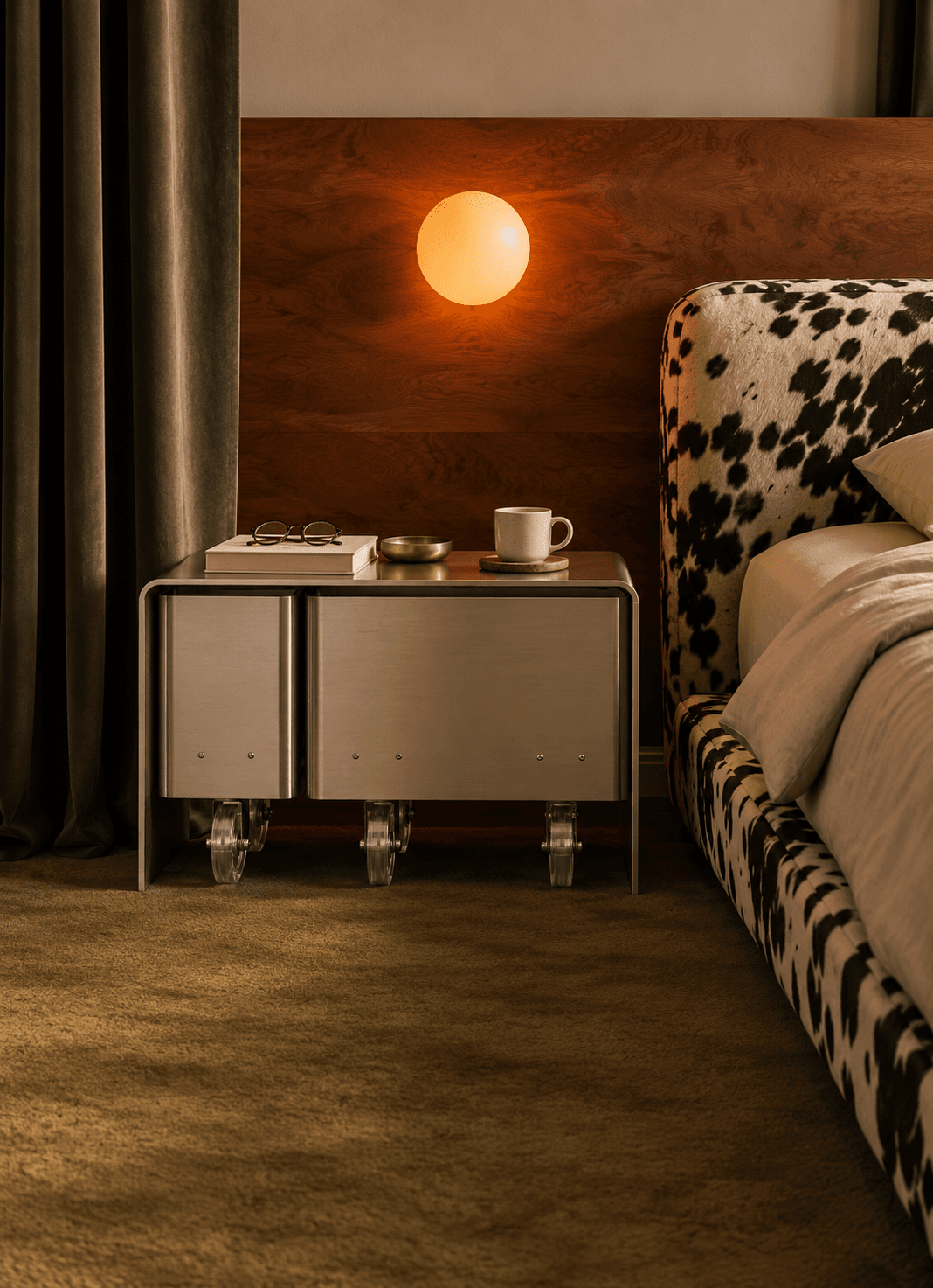The bedside cart at night beneath a glowing globe light, with a book, glasses and a mug on top, beside a bed in patterned upholstery